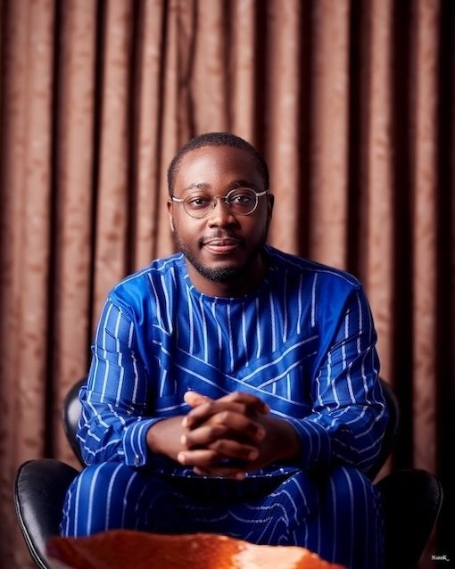 A photo of Osei Boateng, the 2025 Global Citizen Waislitz Award winner and Founder of OKB Hope Foundation. He is wearing glasses and a traditional blue and white striped clothing. Sitting on a black chair in a well-lit room with brown curtains.