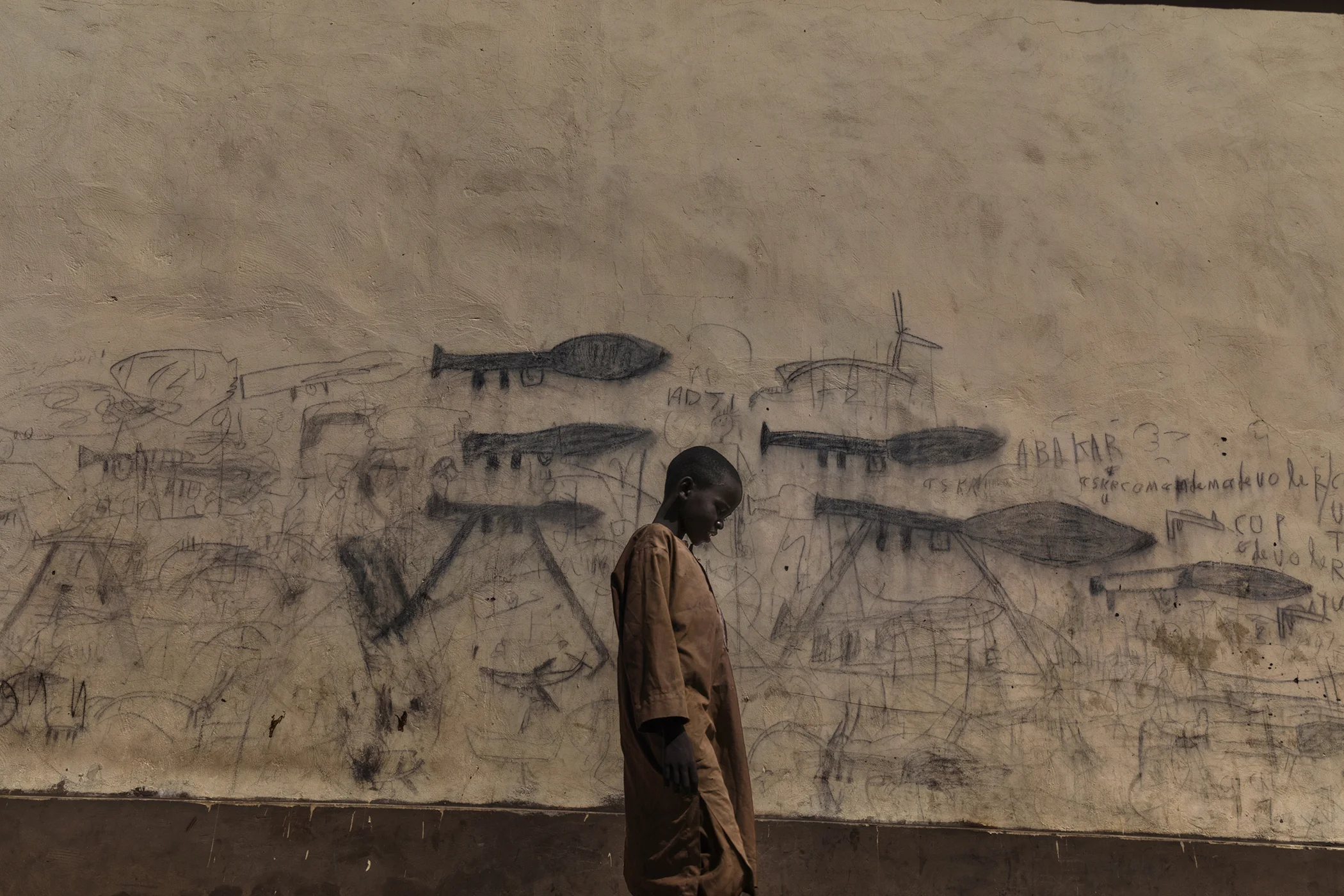 An orphaned boy walks past a wall with drawings depicting rocket-propelled grenade launchers, in Bol, Chad. A humanitarian crisis is underway in the Chad Basin, caused by a complex combination of political conflict and environmental factors.