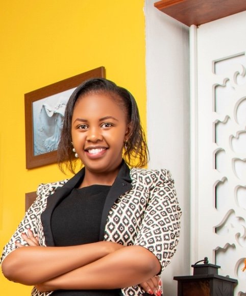 A photo of Maryanne Gichanga, she is standing with her arms crossed, wearing a black and white patterned blazer and a black top, and is smiling. The background  features a yellow wall with photo frames and a patterned white door.