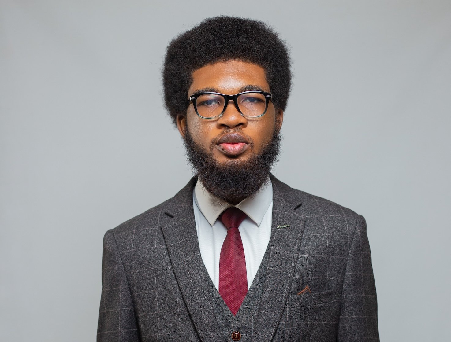 A photo of Joshua Ichor, CEO of Geotek Water Solutions, wearing a black-framed glasses, a dark grey suit with a white grid pattern, a white shirt, and a dark red tie. He stands against a grey wall with a neutral expression.